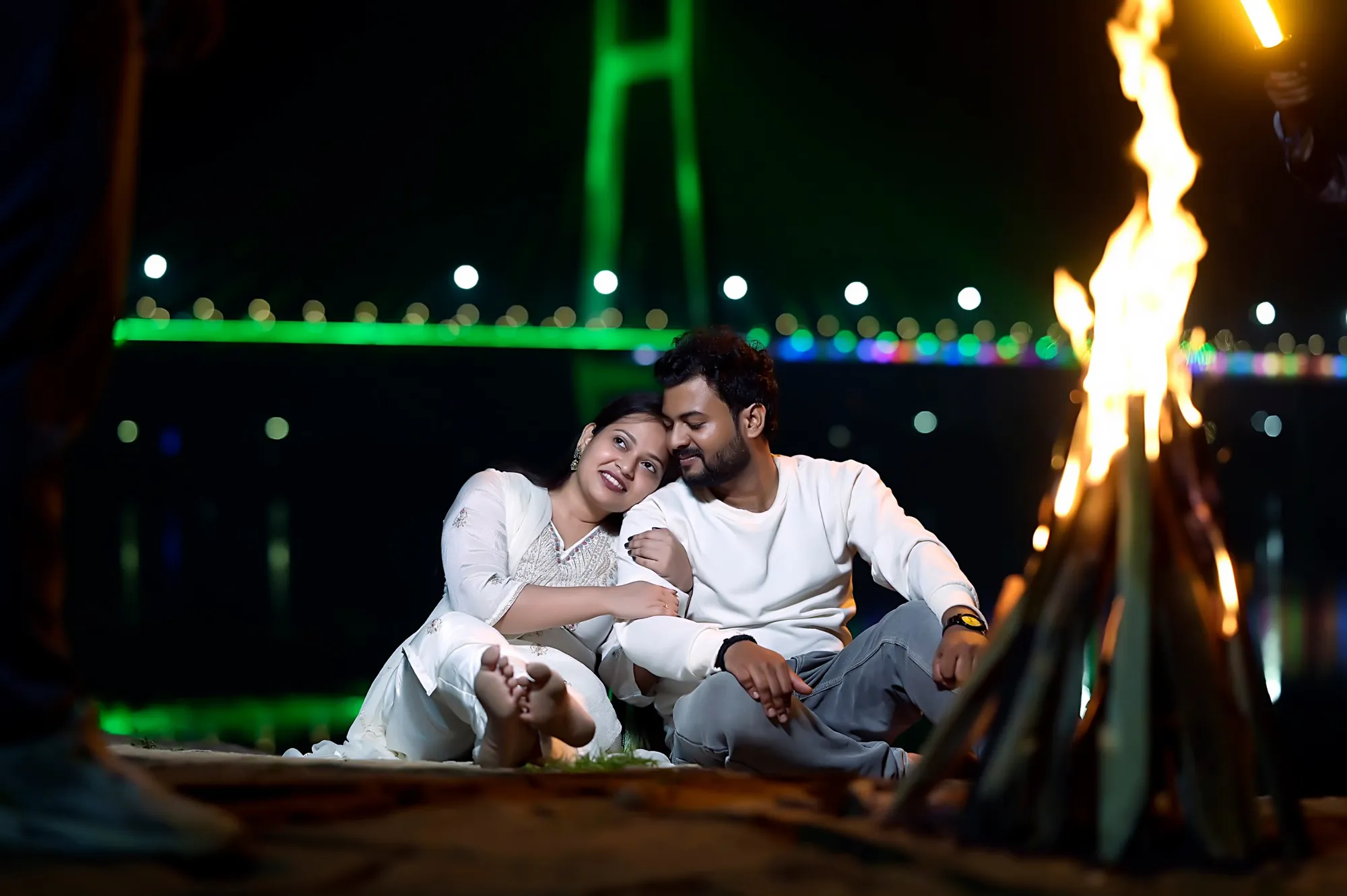 Couple by a bonfire with a lit-up bridge behind them